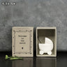 A ceramic figurine of a baby in a stroller placed inside an open brown kraft matchbox with the text 'Welcome to the world' on it, displayed against a concrete background.