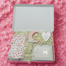 Gift box with baby items including a onesie, blanket, and toys on a pink textured background