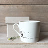 White mug with text and design on a wooden surface with a box and flowers.