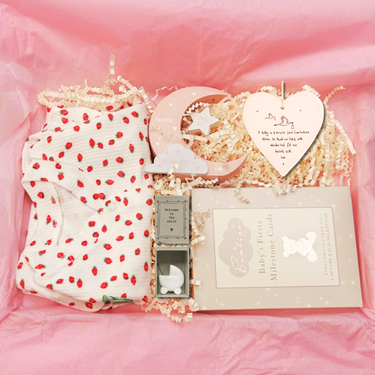 Baby gift set with 100% cotton sleepsuit, heart-shaped plaque, and other items on a pink background