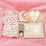 Baby gift set with 100% cotton sleepsuit, heart-shaped plaque, and other items on a pink background